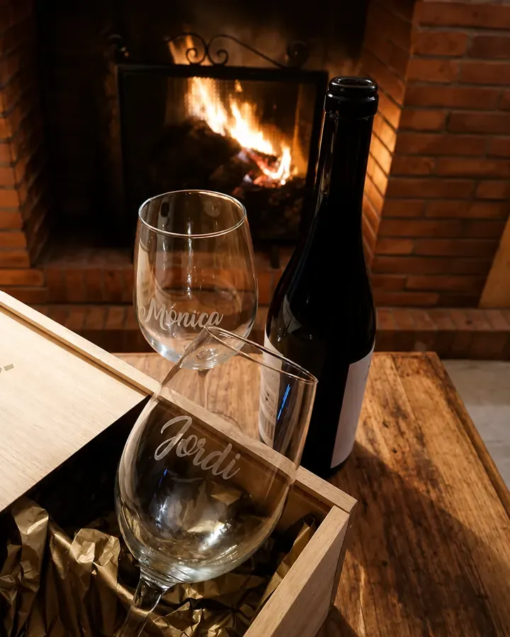 Dos copas de vino grabadas con nombres, junto a una botella y un estuche de madera