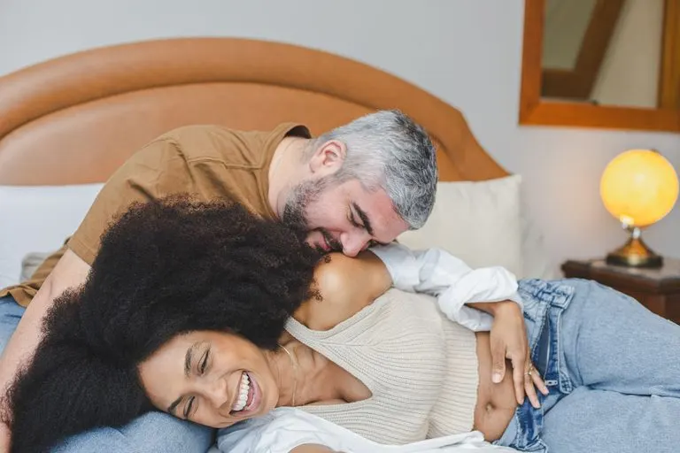 Pareja riendo abrazada entre sábanas blancas, así es Géminis en la cama