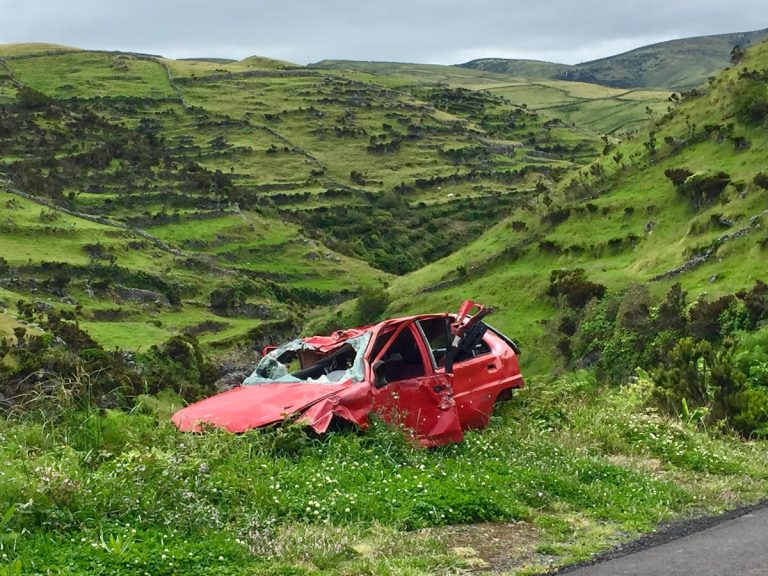 A red wrecked car lies abandoned in a scenic green valley, highlighting rural decay.